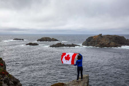 Adventurous woman holding a Canadian Flag on a Rocky Atlantic Ocean Coast during a cloudy day. Taken in Sleepy Cove, Crow Head, Twillingate, Newfoundland, Canada.の写真素材