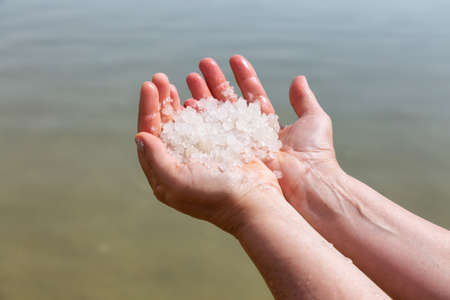 Middle age woman holding salt crystals in her hand with the Dead Sea in the Background during a bright sunny day. Taken in Ein Bokek, Israel.の写真素材