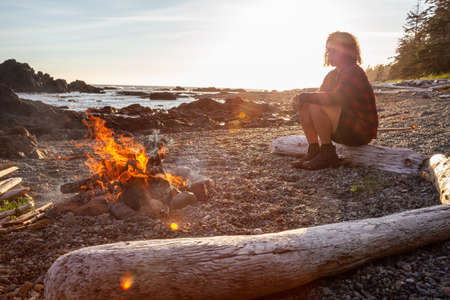 Girl enjoying a camp fire on the beach during a vibrant summer sunset. Taken in Northern Vancouver Island Ocean Coast, BC, Canada.の写真素材