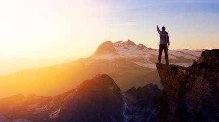 Composite. Adventurous Man Hiker With Hands Up on top of a Steep Rocky Cliff. Sunset or Sunrise. Landscape Taken from British Columbia, Canada. Concept: Adventure, Explore, Hike, Lifestyleの写真素材