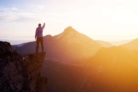 Adventurous Man Hiker With Hands Up on top of a Steep Rocky Cliff. Sunset or Sunrise. Composite. Landscape Taken from British Columbia, Canada. Concept: Adventure, Explore, Hike, Lifestyleの写真素材