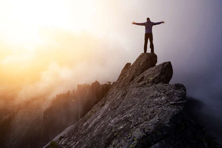 Adventurous Man Hiker With Hands Up on top of a Steep Rocky Cliff. Sunset or Sunrise. Top of Crown Mountain, North Vancouver, British Columbia, Canada. Concept: Adventure, Explore, Hike, Lifestyleの写真素材