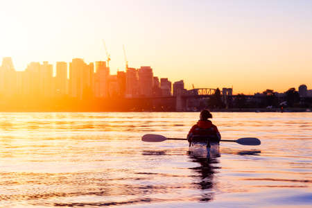 Adventurous Girl Sea Kayaking during a vibrant winter sunrise with City Skyline in Background. Taken in Downtown Vancouver, British Columbia, Canada. Concept: Adventure, Holiday, Vacation, Sportの写真素材