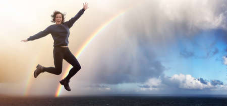 Happy and Excited Girl Jumping with Joy. Ocean Background with Rainbow and Rain Clouds.の写真素材