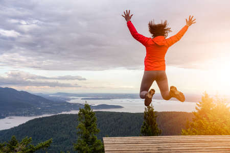 Adventurous Girl Jumping with Joy on top of a mountain with a beautiful Canadian Nature Background. Taken in Bowen Island, near Vancouver, British Columbia, Canada.の写真素材