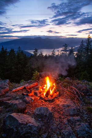 Warm Camp Fire on top of a mountain with Beautiful Canadian Nature Landscape in background during a colorful Sunset. Taken on Bowen Island, near Vancouver, British Columbia, Canada.の写真素材