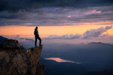 Fantasy Adventure Composite with a Man on top of a Mountain Cliff with Dramatic Landscape in Background during Sunset or Sunrise. Landscape from British Columbia, Canada.の写真素材