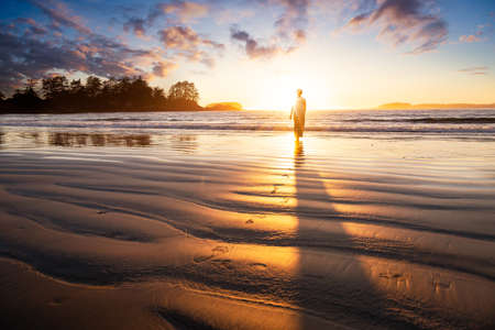 Beautiful woman in a casual dress is walking barefoot on a sandy ocean shore towards the water. Chesterman Beach, Tofino, Vancouver Island, BC, Canada, during golden summer sunset.の写真素材
