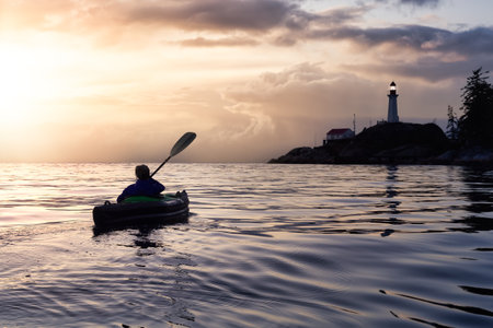 Adventurous woman is sea kayaking near a lighthouse during winter. Dramatic Colorful Sunset Artistic...