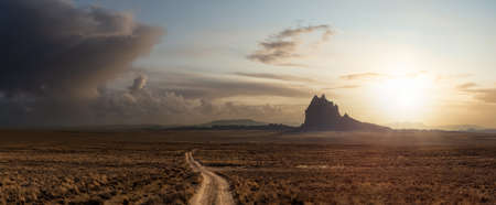 Striking panoramic landscape view of a dirt road in the dry desert with a mountain peak in the background. Colorful Sunset Sky Art Render. Taken at Shiprock, New Mexico, United States.の写真素材