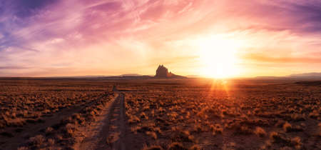 Striking panoramic landscape view of a dirt road in the dry desert with a mountain peak in the background. Colorful Sunset Sky Art Render. Taken at Shiprock, New Mexico, United States.の写真素材