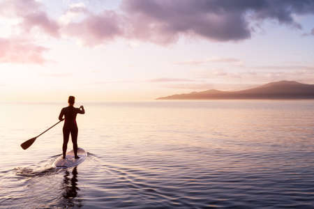 Adventurous woman on a paddle board is paddeling in the Pacific West Coast Ocean. Sunset Sky Art Render. Taken near Spanish Banks, Vancouver, British Columbia, Canada.の写真素材