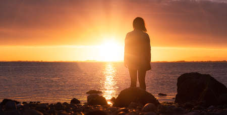 Adventure Caucasian Adult Woman Standing on a rocky beach on the West Coast Pacific Ocean watching a Dramatic Sunset. White Rock, Greater Vancouver, British Columbia, Canada.の写真素材