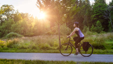 Adventurous White Caucasian Adult Woman riding a road bicycle on a bike path at the famous Stanley Park in a modern city. Sunny Summer Sunset. Downtown Vancouver, British Columbia, Canada.の写真素材