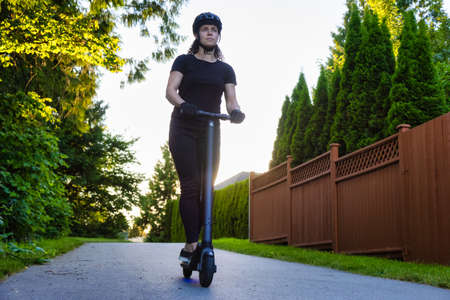 Caucasian Adult Woman riding an electric scooter on a path in a quite neighborhood in a modern city....
