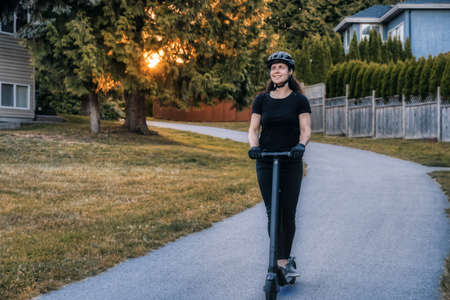 Caucasian Adult Woman riding an electric scooter on a path in a quite neighborhood in a modern city. Taken in Fraser Heights, Surrey, Vancouver, British Columbia, Canada.の写真素材
