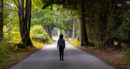 Adventurous Caucasian Woman walking on a Scenic Road by the Vibrant Green Canadian Rainforest. Sunny Summer Morning. Salt Spring Island, British Columbia, Canada.の写真素材