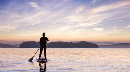 Adventurous Caucasian Adult Woman on a Stand Up Paddle Board is paddling on the West Coast of Pacific Ocean. Sunny Sunrise Sky Art Render. Victoria, Vancouver Island, BC, Canada.の写真素材