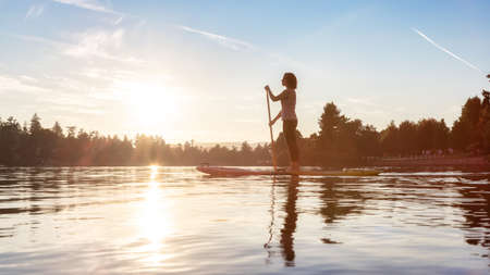 Adventurous Caucasian Adult Woman Paddling on a Stand up Paddle Board in water at a city park. Sunny Sunset Sky. Gorge Park, Victoria, Vancouver Island, BC, Canada.の写真素材