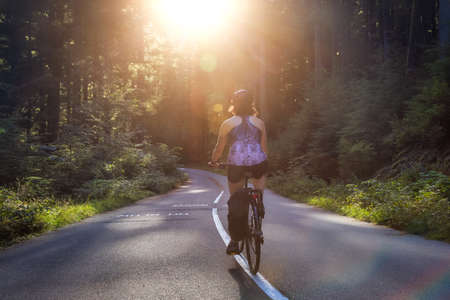 Adventurous White Caucasian Woman on a bicycle riding on a path in Green Canadian Rain Forest. Sunny Sunset. Seymour Valley Trailway in North Vancouver, British Columbia, Canada.の写真素材