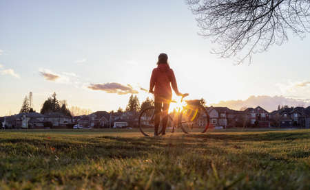 Adventurous Woman Standing with a bicycle at a Park in Modery City Suburbs. Sunset Sky. Fraser Heights, Surrey, Vancouver, British Columbia, Canada.の写真素材