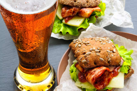 Glass of beer and two burger-like sandwiches with lettuce, bacon, cheese, ketchup on paper, plates and black slate stoneの写真素材
