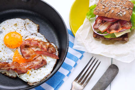 Breakfast: fried eggs and bacon in metal pan and sandwich on white wooden table backgroundの写真素材