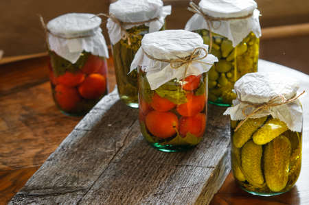Clear glass jars of colorful pickled vegetables: tomatoes and cucumbers in rustic styleの写真素材