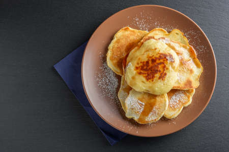 Top view: small pancakes with sugar powder and syrup on brown plate over black slate stone. Copy...