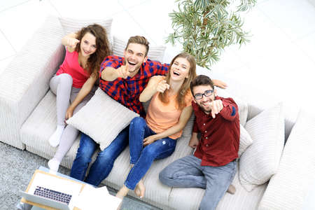 group of laughing friends sitting on sofa and showing forefinger on the camera.の写真素材