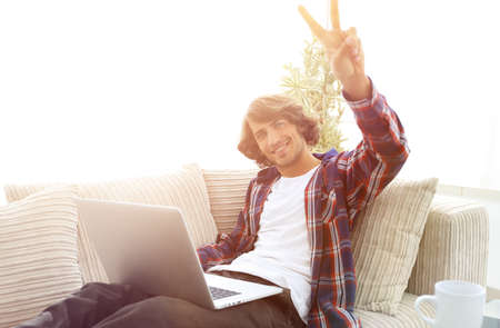 happy guy with laptop sitting on sofa and showing his hand a winの写真素材
