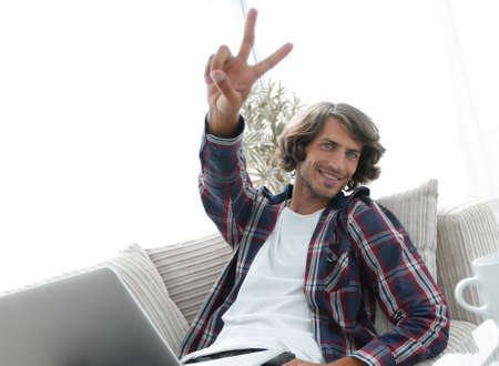 happy guy with laptop sitting on sofa and showing his hand a winning gestureの写真素材