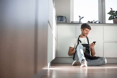 young man with smartphone sitting on kitchen floorの写真素材