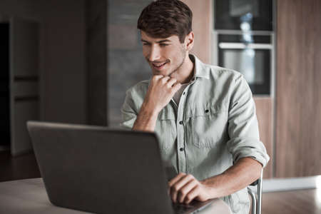 young man sitting in front of open laptop in kitchenの写真素材