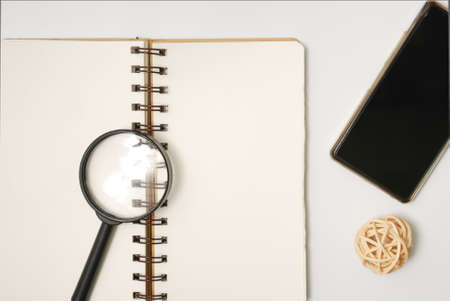 White office desk table with open note book, magnifying glass, rattan ball and smartphone. Top view with copy space, flat lay.の写真素材