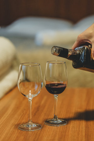 Woman pouring red wine into wine glasses on wooden table in hotel roomの写真素材