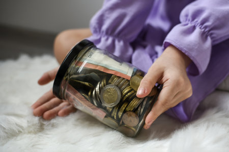 Young woman with money in glass jar on carpet at home, closeup