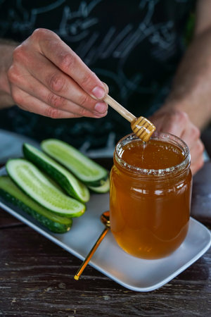 Honey dripping from a wooden honey dipper into a glass jar with cucumber slicesの写真素材
