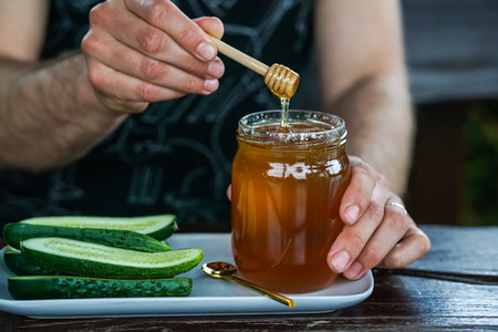 Honey dripping from a wooden honey dipper into a glass jar with cucumber slices on the tableの写真素材
