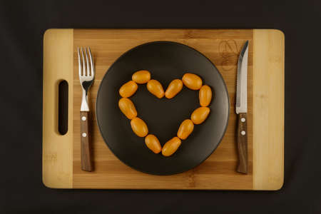 Love of tomatoes. Yellow plum tomatoes arranged on a gray plate in the shape of a heart. Wooden board at the bottom. Next to the knife and fork. Dark background. For gourmets.の写真素材