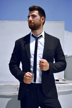Attractive Spanish looking young man with a beard and dark hair and brown eyes is wearing a black suit and tie with a white shirt.の写真素材
