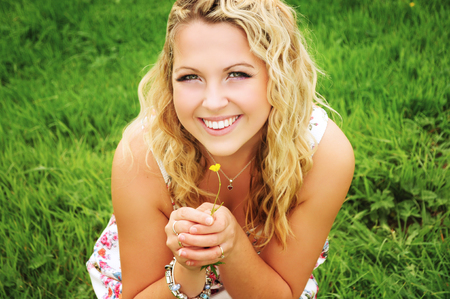 Smiling beautiful blonde woman with curly long hair and natural make-up is sitting in the grass and holding a small yellow flower in her hands.の写真素材