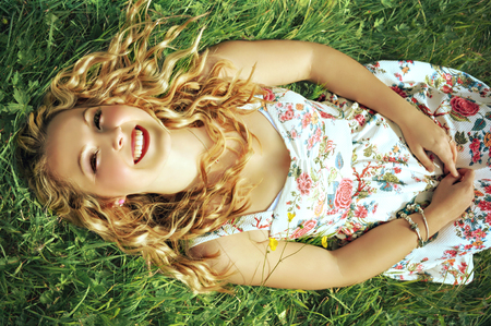 Smiling blonde young woman with curly hair wearing a flower print summer dress with straps and red lipstick is lying in the grass and looking up.の写真素材