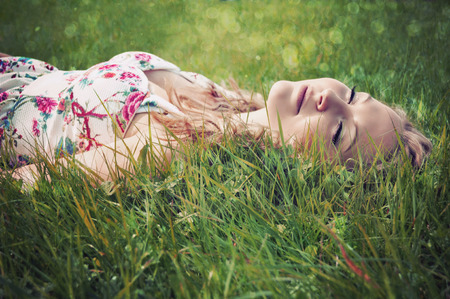 Blonde young woman with curly hair wearing a flower print summer dress with straps is lying down in the grass with her eyes closed.の写真素材