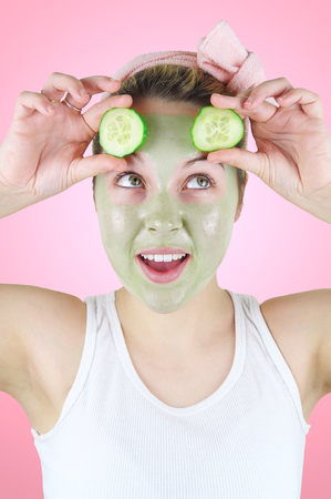 Young funny woman wearing a green facial mask and a pink headband is holding two slices of cucumber above her eyes on pink background.の写真素材