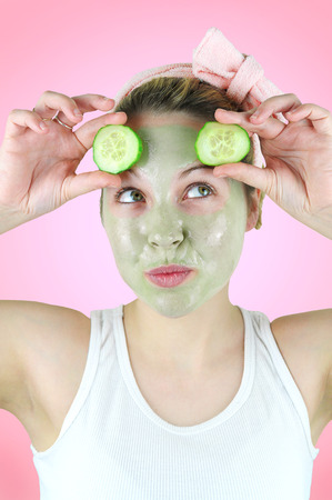 Young funny woman wearing a green facial mask and a pink headband is holding two slices of cucumber above her eyes on pink background.の写真素材