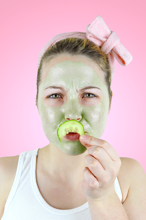 Young funny woman wearing a green facial mask and a pink headband is holding a bitten cucumber slice in front of her lips on pink background.の写真素材