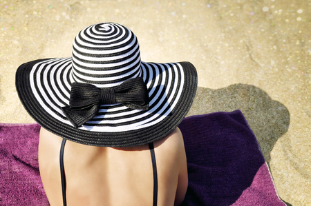 Elegant woman wearing a black bikini and a large black and white striped summer hat is lying comfortably on the towel on the beach.の写真素材