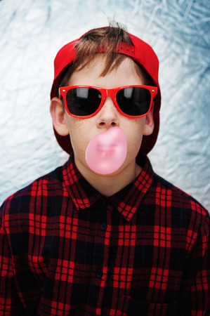 Funky teenage boy wearing red sunglasses, red baseball cap and red checked shirt is blowing pink bubbles with his chewing gum.の写真素材