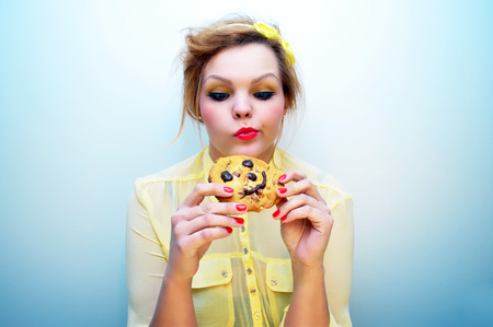 Young attractive woman with red hair and a yellow bow headband wearing a yellow chiffon blouse is holding a smiling chocolate chip cookie and looking at it with an indecisive look.の写真素材